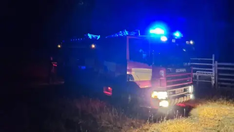 A fire engine at night parked up in a field. The vehicle's blue lights are flashing and it is only illuminated by its headlights. It is predominately red with yellow high viz squares down its side. 