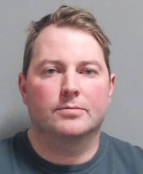 PSNI Jonathan Creswell has short brown hair parted to the side and light blue eyes. He is wearing a blue/grey t-shirt and is staring straight ahead in a police mugshot. 