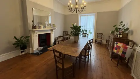 A room with grey walls and a wooden floor with a large table in the centre surrounded by chairs, a white fireplace and plants in pots in various places