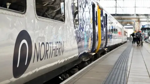 Northern A set of carriages on a Northern train running along a platform with the company sign seen on the outside of one of the cars. People can be seen at the end of platform waiting to alight. 