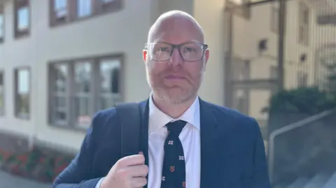 Rob Curgenven looks at the camera with a neutral expression. He is wearing glasses, a blue suit and a patterned tie. He has a rucksack over one shoulder which he is holding with one hand.