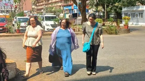 Mukesh Kumar Three women walking across a road in Goa, all have long brown hair, woman on left is wearing cream blouse and brown skirt with sandles, woman in middle is wearing blue dress with purple scarf, woman on right is in checkered black and white top with black trousers, a bright blue crossbody bag and sandles

There are plants and vehicles visible in the background