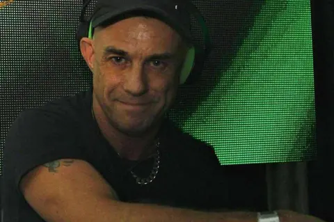 Daz Willott A man in a black baseball cap and T-shirt has headphones on one ear and a silver chain round his neck. He is looking at the camera as he stands in front of a screen showing a green patterned background.