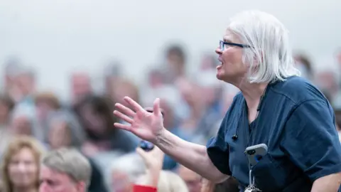 Woman wearing glasses raises an arm as she shouts during a town hall meeting.