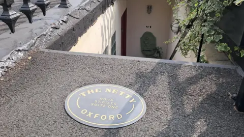 A photo of steps leading down from street level, with a plaque on the ground reading "The Netty - Oxford".