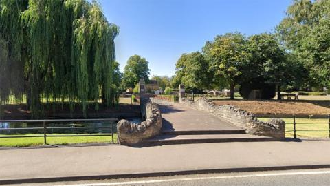 A small bridge over a waterway with trees either side, leading to the gates of the park