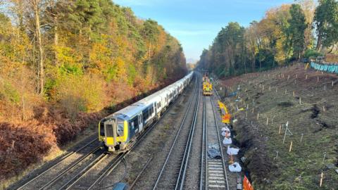 Image shows a train travelling alongside a slope which is having emergency works done to it to prevent a landlsip