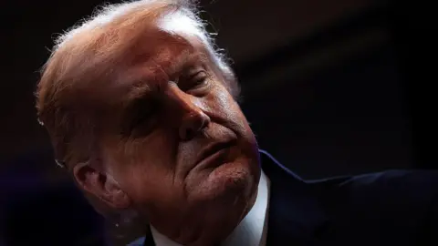 Trump looks away. He is stood in front of a dark background, wearing a suit. 