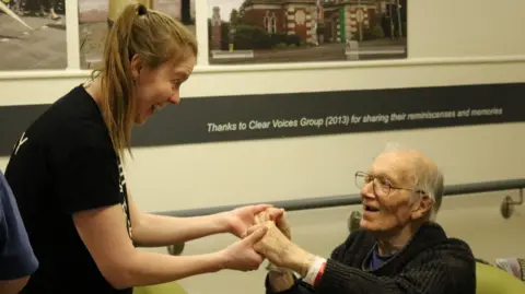 Bedfordshire Hospitals NHS Foundation Trust Jenny Howells, wearing a black T-shirt, smiles and holds the hands of a man who is sitting down. He is smiling back at her.