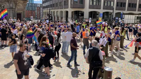 Dozens of people are gathered together, many holding large or small rainbow flags. There is rainbow confetti on the floor