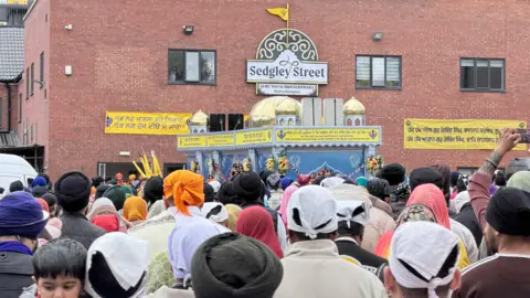 Jules Singh Image shows a Sikh temple, a large red brick building with a large sign bearing it's name: Sedgley Street - in front of the building are hundreds of people, the backs of their heads are facing the camera and they are wearing turbans of all colours and scarfs.