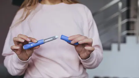 An anonymous woman in a pink top holding a weight loss jab pen in one hand and the cap of the pen in the other. Stairs in the background.