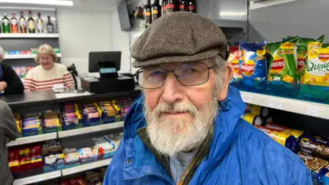 John Devine/BBC John Dudeney, 80, has a grey bushy beard and moustache, glasses and a flat cap, blue mac with a tartan fleece, blue shirt and white T-shirt underneath. Behind him is a volunteer shop keeper and a range of products on display shelves.