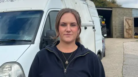 Ceylone Fletcher stands in front of a white horsebox van, with part of the word 'Horses' visible on the top. She is wearing a dark sweatshirt and stood in a rural setting, with a stable behind her and a part of a tractor visible. The sky is partly cloudy, and there are trees in the distance.