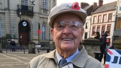 Jack Waterfield in a town centre dressed in a beige military cap with a poppy sewn to the front. He is wearing glasses and smiles and the camera. He is also wearing a blue and white striped shirt with a dark blue tie and over the top is a beige collared coat. 