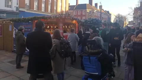 A shot of the Christmas Market in York, with a large fir tree archway, decorated with gold, green and red baubles and illuminated stars. A mixed crowd of people can be seen walking beneath it.