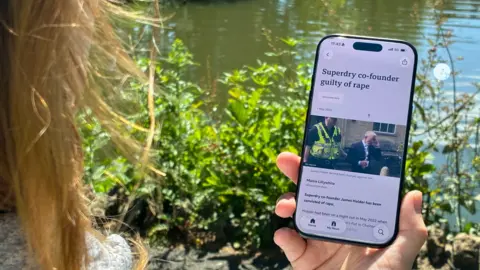 A woman looks at a BBC news story on a mobile phone. You can only see her hair and hand. The headline reads: Superdry co-founder guilty of rape.