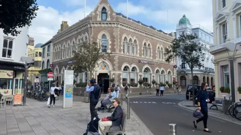 A street corner in a town centre with a large historic brick building featuring arched windows and a central entrance. People are walking, sitting on benches, cycling, and crossing the road, with shops, bicycles, trees, and surrounding older buildings visible under a partly cloudy sky.