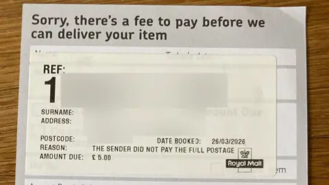 Supplied A leaflet from the Royal Mail saying "Sorry, there's a fee to pay before we can deliver your item". There is a large reference number, but the surname and address of the recipient have been blurred out. The date on the leaflet is 26 March 2026. It also says "the sender did not pay the full postage" and that the "amount due" is £5.