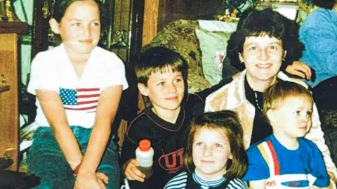 Family photo A woman sits on a chair surrounded by her four children. The woman is smiling at the camera. The four children are all looking off camera. The youngest sits on her knee.