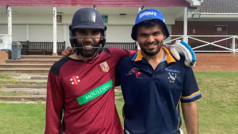 BBC Mohammed on the left and Yash on the right on the pitch at Slough Cricket Club posing for the camera with their arms around each other's shoulder. Mohammed is wearing a navy blue cricket helmet and has a dark beard and has a red cricketing top on as well as a white batting glove on his left hand. Yash has a blue bucket hat on that says "London spirit" on it and is wearing a navy blue polo top. Behind them you can see the front of the cricket pavilion which has some brick steps up to it and some brown benches in front of white shutters.