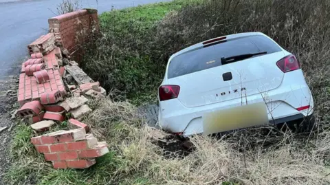 A white Seat Ibiza in a brook. The bridge's brick wall next to it is damaged.