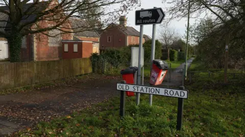 A concrete footpath running diagonally from left to right into the distance. There  are brown leaves on the path and houses to the left of the footpath. There is a brown All Routes sign and two red dog poo bins. A white street sign in centre states Old Station Road in black letters.  