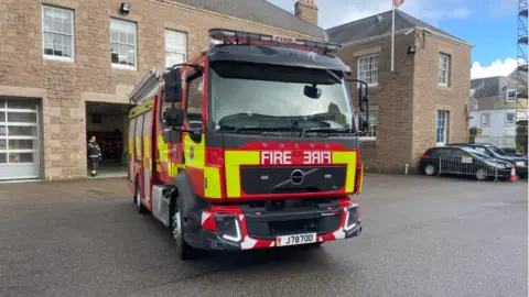 A large fire vehicle parked in front of a large brown building - the fire station. Normal cars can be seen parked to the right.