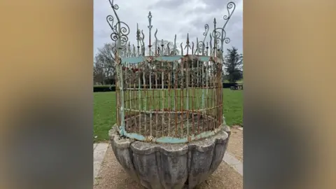 An out of use fountain, which is made up of a rusty, light blue metal top and a concrete base. Behind it there is a field