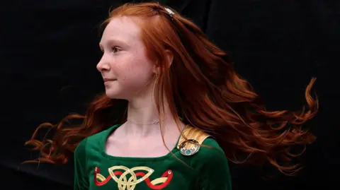 A young girl with red hair, wearing a green velvet Irish dancing outfit