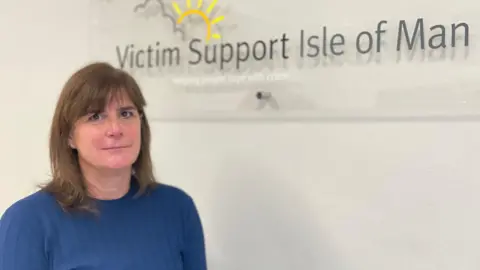 Lorna Trevethan, who has short brown hair and is wearing a blue jumper. She is standing in front of a sign that reads Victim Support Isle of Man.