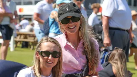 Pacemaker Two girls are smiling at the camera. They both have long blonde hair an are wearing black sunglasses. The girl on the left is wearing a white top and the girl on the right is wearing a pink shirt and black cap.