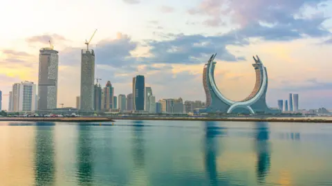 Getty Images A waterside view of Lusail, Qatar. Across a stretch of blue water, skyscrapers can be seen, including an unusually shaped horseshoe skyscraper-type building.