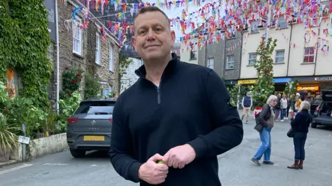 A man in a black shirt stands in the streets of Padstow under a may pole.