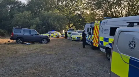 Norfolk Constabulary Seven police vehicles are parked on a road, blocking it off entirely. They are a mix of police cars and riot vans. There are two officers, dressed in black, standing next to the vehicles. To the left, further on the grass, there is a dark grey off-road vehicle.
