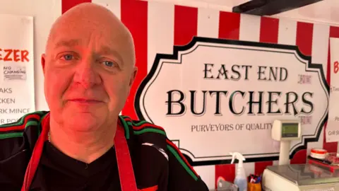 Ben Parker/BBC A man wearing a black Arsenal FC top with a red apron, behind him is a sign that says East End Butchers.