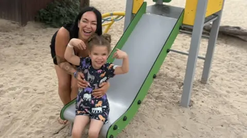 Melanie Prince A close of Melanie Prince's daughter Mabel, who is playing on a slide in a sand pit. Melanie is holding her by the waist. 
