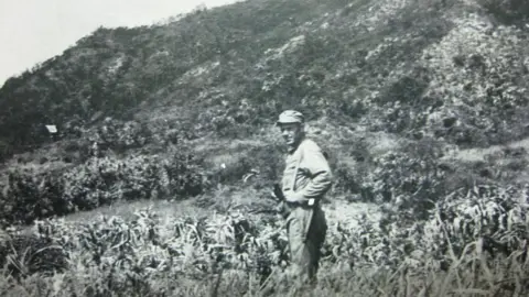 Trudy Johnson A black and white image of George Clark standing on Aka island in front of a cliff. To one side, a row of troops carrying a white flag is just visible 