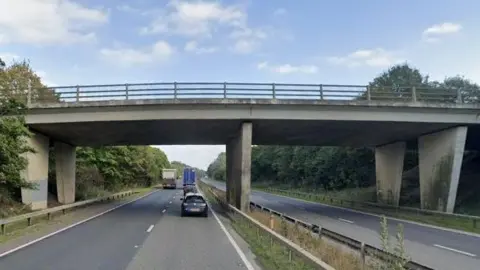 Two-lane motorway, showing concrete road bridge with two wide pillars on either side and one in the centre. There is a railing on the edge of the road crossing the bridge. There are trees and hedges either side of the bridge.There is traffic heading away from the camera including cars and lorries.