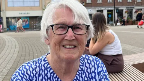 Barbara Logan is sitting on a bench in a square in Truro. She is wearing a blue and white patterned top and a pair of glasses. She has short white hair and is smiling. A woman with long dark blonde hair is sitting behind her. There are a few people walking across the square in the background and there is a Primark, a JD Sports and a Wetherspoons.