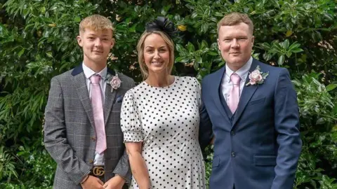Supplied A woman stands in the middle of a teenage boy and man wearing a black and white polka dot dress and black flower headband smiling with blonde hair mostly tied back. A teenage boy is wearing a tweed suit next to her with a rose broach and pink tie. A man to the right is wearing a navy suit, pink tie and rose broach. They are stood in front of a bush.