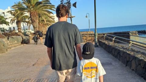 Dad Liam walking along a promenade next to the sea with his adoptive son. There are palm trees in the background and a woman pushing a bike