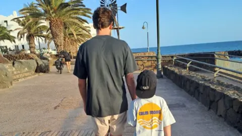 BBC Dad Liam walking along a promenade next to the sea with his adoptive son. There are palm trees in the background and a woman pushing a bike