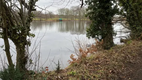 A view of Poynton Pool. It is taken from one of the embankments and shows trees on the side and looking across the lake.