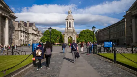 Getty Images People walk with their backs to the camera through Trinity College Dublin's campus. The campus is paved and has some grass areas. It is surrounded by large old grey buildings. The centre of the campus has a large tower.