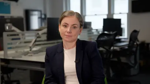 Ben Moore/ BBC Woman looks at camera in an office. She has blonde hair, and is wearing a white t-shirt and a black suit.