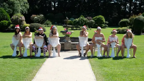 Deafblind UK A group of women are sitting on a row of chairs, naked, while wearing santa hats. Their private parts are covered by a white material. They are sat in a large green garden infront of a fountain.