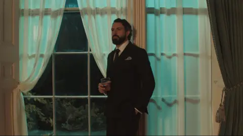 WorldProductions/PA Tom Cullen is wearing a dark suit, pocket tie and a tie. He's standing in front of a large window, with curtains either side, holding a glass of a drink.
