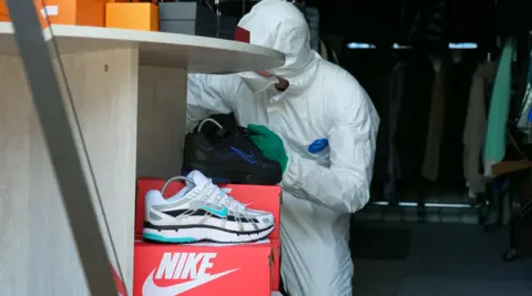 Man in white suit, face covering and disposable gloves examines a product. He rests on one knee looking closely at something. A white counterfeit 'Nike' trainer and red shoebox are at the front of the photo