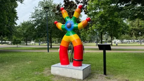 Andrew Turner/BBC A four-armed, two legged sculpture painted in concentric bands (from outer to inner) red, orange, yellow, green, blue and purple. The design has a ball in the middle and on the tips of all four arms.